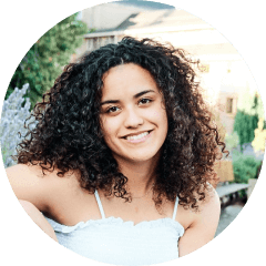 A person with curly hair smiles outdoors, wearing a light-colored top. There are plants and a building in the background.