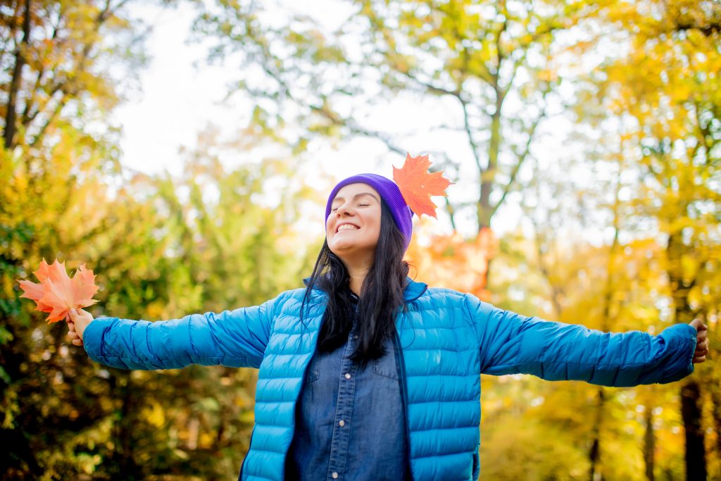 A person wearing a blue jacket and purple beanie smiles with eyes closed in a forest, holding autumn leaves in both hands—a perfect Sample Post #1 for capturing the cozy essence of fall.